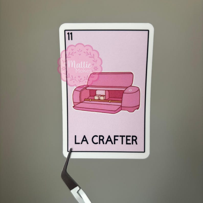 以下が含まれることがあります： ピンク色のクラフトマシンのイラストと「LA CRAFTER」の文字が入ったピンク色のステッカー。左上に数字の11があります。ピンセットでステッカーを持っています。