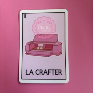以下が含まれることがあります： 「LA CRAFTER」の文字が入ったピンク色のクラフトマシンが描かれたピンク色のステッカー。ステッカーには白い縁取りがあり、左上隅には数字の「11」が入っています。ステッカーには、装飾的なフォントで「Mattie Makes」という文字も入っています。