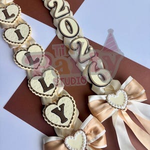 May include: Cream-colored graduation leis with gold and white ribbons, heart-shaped accents, and the letters "L", "Y", and the year "2026". The leis feature bows and a graduation cap.