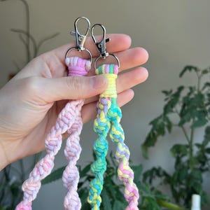 Puede incluir: Dos llaveros de crochet coloridos con cierres plateados. Un llavero es rosa y blanco, el otro es verde, amarillo y morado.