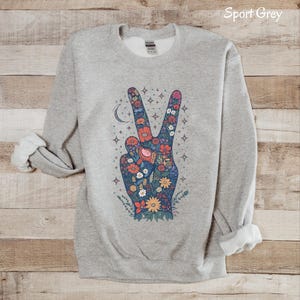 May include: Sport Grey sweatshirt with a floral peace sign graphic. The peace sign is filled with colorful flowers, a crescent moon, and stars. The sweatshirt has a crew neck and long sleeves with rolled-up cuffs.
