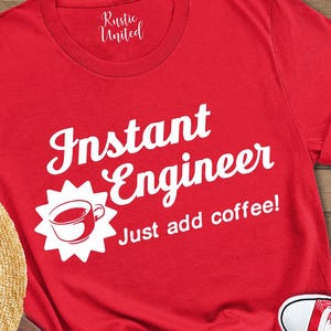 May include: A red t-shirt with the text "Instant Engineer Just add coffee!" is displayed with a straw hat, brown backpack, blue jeans, and red sneakers. The items are arranged on a wooden surface.