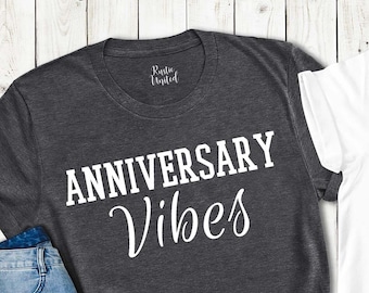 May include: Two t-shirts, one charcoal gray crew neck and one white V-neck, both with the text "ANNIVERSARY Vibes" in white and black, respectively. A pair of blue jeans and sunglasses are next to the gray shirt. The shirts are from Joy Apparel USA.
