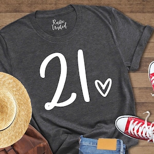 K&ouml;nnte beinhalten: Dunkelgraues meliertes T-Shirt mit der Zahl 21 und einem Herz-Grafik. Das Bild zeigt auch rote Turnschuhe, einen Strohhut, einen braunen Rucksack und blaue Jeans, die auf einer Holzoberfl&auml;che angeordnet sind.
