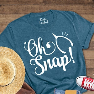 May include: A flat lay featuring a teal t-shirt with the text "Oh Snap!", a straw hat, a brown leather backpack, blue jeans, and red sneakers. The items are arranged on a wooden surface, creating a casual, lifestyle aesthetic.