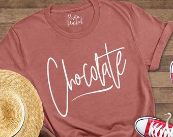May include: A heather mauve t-shirt with the word "Chocolate" in white script. The image includes a straw hat, brown backpack, blue jeans, and red sneakers. The items are arranged on a wooden surface.