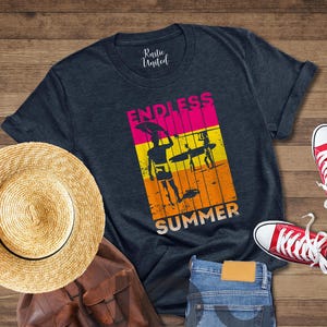 May include: A navy blue t-shirt with the words "ENDLESS SUMMER" and a graphic of surfers. The image also includes a straw hat, a brown backpack, blue jeans, and red sneakers.