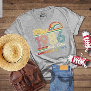 May include: A light grey t-shirt with a vintage 1986 graphic, a straw hat, a brown leather backpack, blue jeans, and red sneakers. The shirt has a rainbow design and the words "Vintage 1986 Limited Edition Original Parts."