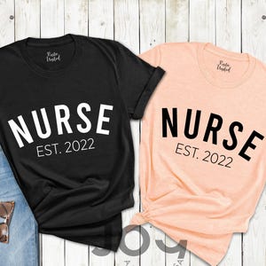 May include: Black and peach-colored t-shirts with the word "NURSE" in large white and black letters, respectively. The text "EST. 2022" is below the word "NURSE". A pair of blue jeans and sunglasses are next to the black shirt.