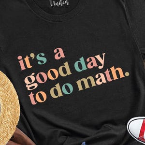May include: A black t-shirt with the text "it's a good day to do math" in colorful letters, paired with red sneakers, blue jeans, a straw hat, and a brown backpack. The items are arranged on a wooden surface.