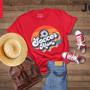 May include: A red t-shirt with a soccer ball graphic and the words "Soccer Mom" in a retro design. The image includes a straw hat, brown backpack, blue jeans, and red trainers. The items are arranged on a wooden surface.