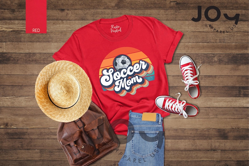 May include: A red t-shirt with a soccer ball graphic and the words "Soccer Mom" in a retro design. The image includes a straw hat, brown backpack, blue jeans, and red trainers. The items are arranged on a wooden surface.