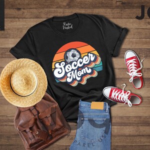 May include: Black t-shirt with a "Soccer Mom" graphic, a straw hat, a brown backpack, blue jeans, and red trainers. The shirt features a soccer ball design with a retro colour scheme.