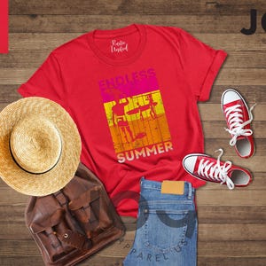 May include: A red t-shirt with "ENDLESS SUMMER" text and graphic, paired with red sneakers, a straw hat, a brown backpack, and blue jeans. The image is on a wooden surface.