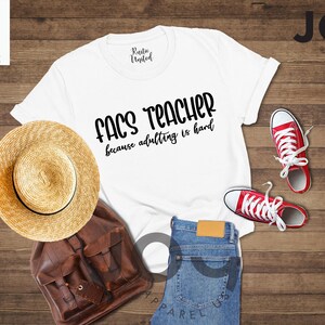 May include: A white t-shirt with the text "FACS TEACHER because adulting is hard" is displayed with a straw hat, brown backpack, blue jeans, and red sneakers. The items are arranged on a wooden surface.