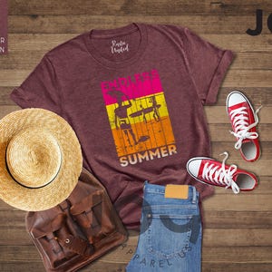 May include: A maroon t-shirt with a graphic that says "Endless Summer" with a surfer theme, red sneakers, a straw hat, a brown backpack, and blue jeans. The items are arranged on a wooden surface.
