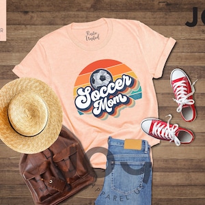 May include: A peach-coloured t-shirt with a retro design that says "Soccer Mom" with a soccer ball graphic. The image also includes red trainers, a straw hat, a brown backpack, and blue jeans.