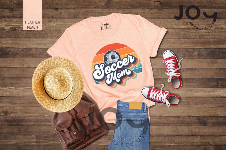 May include: A peach-coloured t-shirt with a retro design that says "Soccer Mom" with a soccer ball graphic. The image also includes red trainers, a straw hat, a brown backpack, and blue jeans.