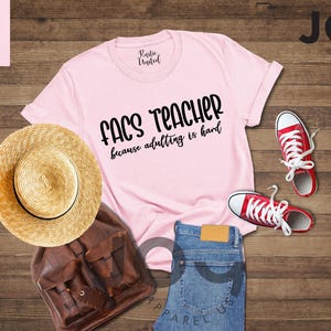 May include: A light pink t-shirt with the text "FACS TEACHER because adulting is hard" in black. The image also includes a straw hat, brown backpack, blue jeans, and red sneakers.
