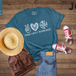 May include: A flat lay featuring a teal t-shirt with the words "Peace, Love, Technology", a straw hat, a brown backpack, blue jeans, and red sneakers. The items are arranged on a wooden surface.