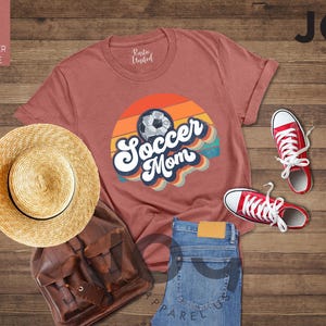 May include: A mauve t-shirt with a retro "Soccer Mom" design and a soccer ball graphic. The image also shows red trainers, a straw hat, a brown backpack, and blue jeans.