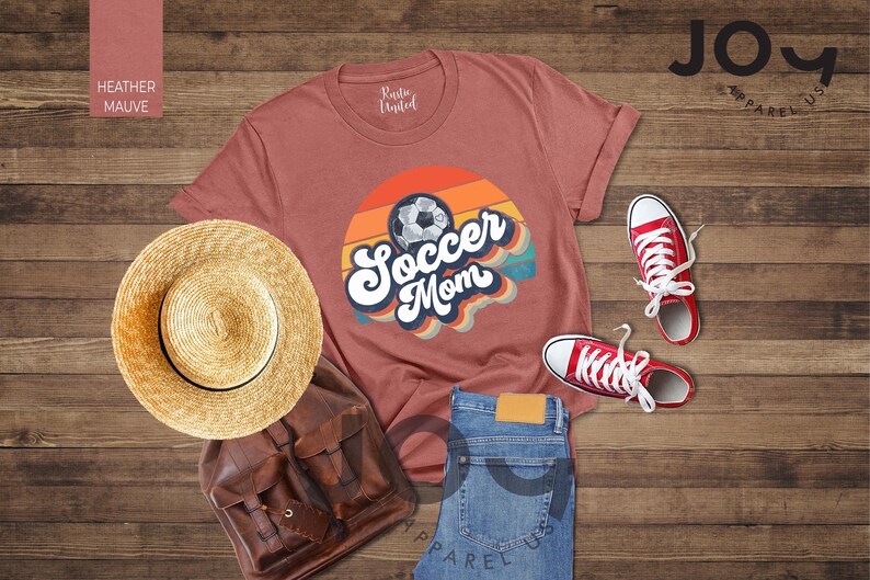 May include: A mauve t-shirt with a retro "Soccer Mom" design and a soccer ball graphic. The image also shows red trainers, a straw hat, a brown backpack, and blue jeans.