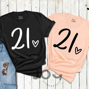 K&ouml;nnte beinhalten: Zwei T-Shirts, eines in Schwarz und eines in Pfirsich, zeigen jeweils die Zahl "21" in einer gro&szlig;en, stilisierten Schriftart mit einem Herzsymbol. Das schwarze Shirt ist mit einer Jeans und einer Sonnenbrille kombiniert. Die Shirts sind von Joy Apparel USA.