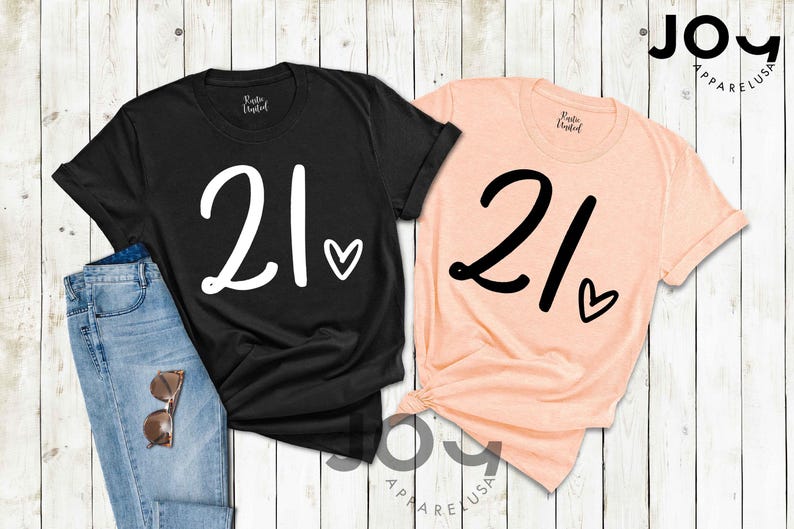 K&ouml;nnte beinhalten: Zwei T-Shirts, eines in Schwarz und eines in Pfirsich, zeigen jeweils die Zahl "21" in einer gro&szlig;en, stilisierten Schriftart mit einem Herzsymbol. Das schwarze Shirt ist mit einer Jeans und einer Sonnenbrille kombiniert. Die Shirts sind von Joy Apparel USA.