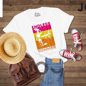 May include: White t-shirt with "ENDLESS SUMMER" graphic, straw hat, brown backpack, blue jeans, and red sneakers. The shirt features a colorful design of a person with a surfboard. The items are arranged on a wooden surface.