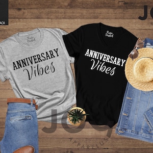 May include: Two t-shirts, one light grey and one black, with the text "ANNIVERSARY Vibes" in a script font. A straw hat, denim jacket, and jeans are also visible. The items are arranged on a wooden surface.