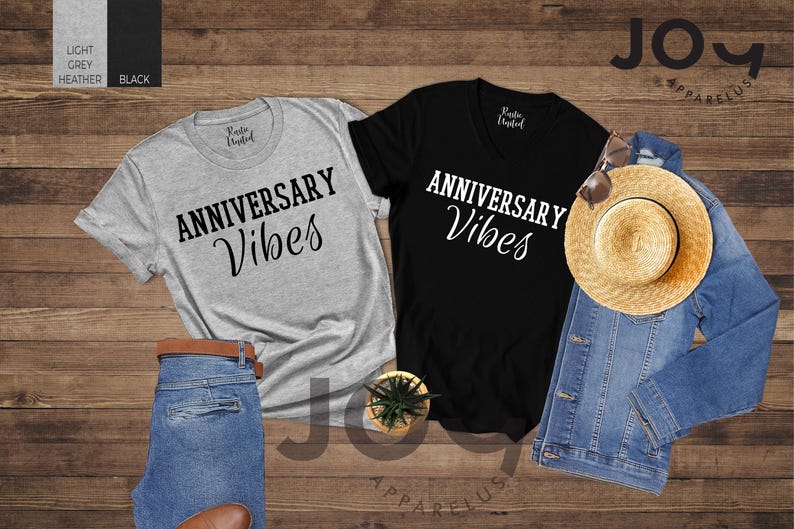 May include: Two t-shirts, one light grey and one black, with the text "ANNIVERSARY Vibes" in a script font. A straw hat, denim jacket, and jeans are also visible. The items are arranged on a wooden surface.