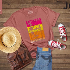 May include: A heather mauve t-shirt with the words "ENDLESS SUMMER" in a colorful graphic, paired with red sneakers, blue jeans, a straw hat, and a brown leather backpack. The items are arranged on a wooden surface.