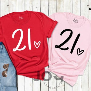 K&ouml;nnte beinhalten: Zwei T-Shirts, eines rot und eines rosa, jeweils mit der Zahl "21" in Schreibschrift und einem kleinen Herz. Das rote Shirt hat wei&szlig;en Text und Herz, w&auml;hrend das rosa Shirt schwarzen Text und Herz hat. Eine Jeans und eine Sonnenbrille sind ebenfalls zu sehen.