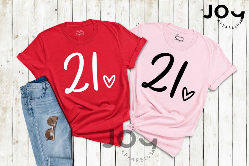 K&ouml;nnte beinhalten: Zwei T-Shirts, eines rot und eines rosa, jeweils mit der Zahl "21" in Schreibschrift und einem kleinen Herz. Das rote Shirt hat wei&szlig;en Text und Herz, w&auml;hrend das rosa Shirt schwarzen Text und Herz hat. Eine Jeans und eine Sonnenbrille sind ebenfalls zu sehen.