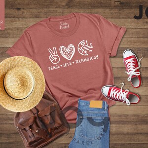 May include: A heather mauve t-shirt with the words "Peace, Love, Technology" in white. The image includes a straw hat, brown backpack, blue jeans, and red sneakers. The items are arranged on a wooden surface.