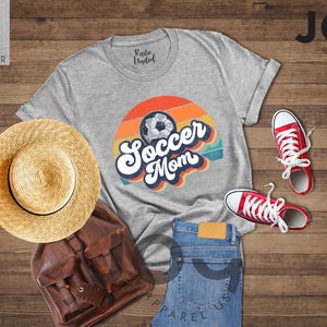May include: A light grey heather t-shirt with a retro design that says "Soccer Mom" with a soccer ball graphic. The image also includes red trainers, blue jeans, a straw hat, and a brown backpack.