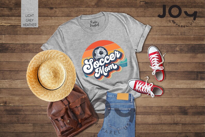 May include: A light grey heather t-shirt with a retro design that says "Soccer Mom" with a soccer ball graphic. The image also includes red trainers, blue jeans, a straw hat, and a brown backpack.