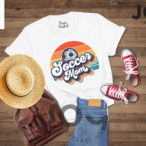 May include: A white t-shirt with a retro "Soccer Mom" design and a soccer ball graphic. The image includes red trainers, blue jeans, a straw hat, and a brown backpack. The shirt is from Joy Apparel US.