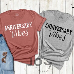 May include: Two t-shirts with the text "ANNIVERSARY Vibes" in white and black. One shirt is a dusty rose color, the other is gray. A pair of blue jeans and sunglasses are next to the dusty rose shirt. The shirts are from Joy Apparel USA.