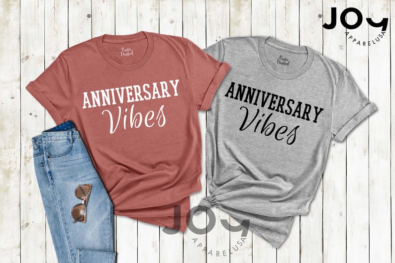 May include: Two t-shirts with the text "ANNIVERSARY Vibes" in white and black. One shirt is a dusty rose color, the other is gray. A pair of blue jeans and sunglasses are next to the dusty rose shirt. The shirts are from Joy Apparel USA.