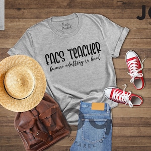 May include: A light grey heather t-shirt with the text "FACS TEACHER because adulting is hard" is displayed with red sneakers, a straw hat, a brown backpack, and blue jeans on a wooden surface.