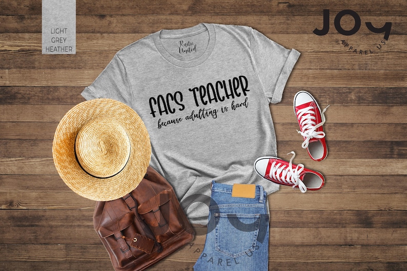 May include: A light grey heather t-shirt with the text "FACS TEACHER because adulting is hard" is displayed with red sneakers, a straw hat, a brown backpack, and blue jeans on a wooden surface.