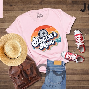 May include: A pink t-shirt with a retro "Soccer Mom" design and a soccer ball graphic. The image also shows red trainers, blue jeans, a straw hat, and a brown backpack.