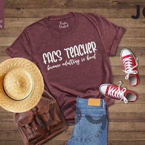 May include: A heather maroon t-shirt with the text "FACS TEACHER because adulting is hard" is displayed with red sneakers, a straw hat, a brown backpack, and blue jeans on a wooden surface. The shirt is from Rustic United.