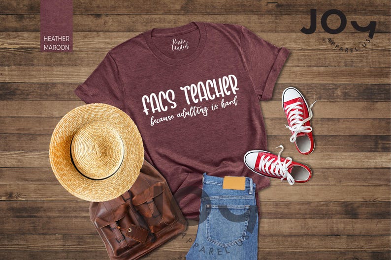 May include: A heather maroon t-shirt with the text "FACS TEACHER because adulting is hard" is displayed with red sneakers, a straw hat, a brown backpack, and blue jeans on a wooden surface. The shirt is from Rustic United.