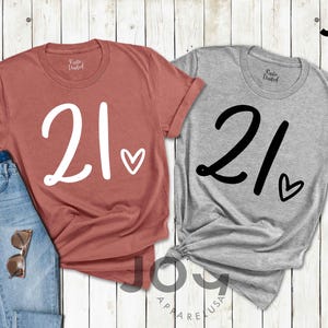 K&ouml;nnte beinhalten: Zwei T-Shirts, eines in Ziegelrot und eines in Grau, jeweils mit der Zahl "21" in Schreibschrift und einem Herzsymbol. Die Shirts werden mit einer Jeans und einer Sonnenbrille auf einer wei&szlig;en Holzoberfl&auml;che pr&auml;sentiert. Der Markenname "JOY APPAREL USA" ist sichtbar.