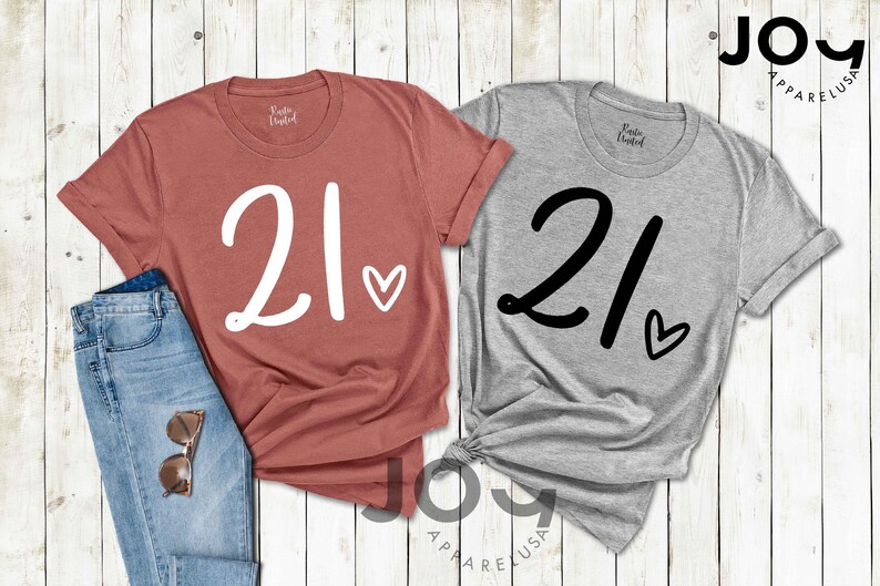 K&ouml;nnte beinhalten: Zwei T-Shirts, eines in Ziegelrot und eines in Grau, jeweils mit der Zahl "21" in Schreibschrift und einem Herzsymbol. Die Shirts werden mit einer Jeans und einer Sonnenbrille auf einer wei&szlig;en Holzoberfl&auml;che pr&auml;sentiert. Der Markenname "JOY APPAREL USA" ist sichtbar.