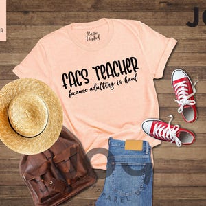 May include: A peach-colored t-shirt with the text "FACS TEACHER because adulting is hard" is displayed with red sneakers, a straw hat, a brown leather backpack, and blue jeans on a wooden surface.
