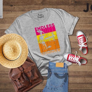 May include: A flat lay featuring a light grey t-shirt with the words "ENDLESS SUMMER" in a colorful graphic. The image includes a straw hat, brown backpack, blue jeans, and red sneakers. The items are arranged on a wooden surface.
