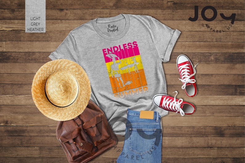 May include: A flat lay featuring a light grey t-shirt with the words "ENDLESS SUMMER" in a colorful graphic. The image includes a straw hat, brown backpack, blue jeans, and red sneakers. The items are arranged on a wooden surface.